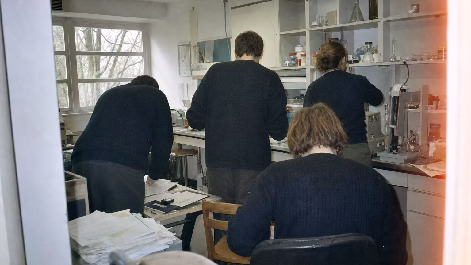Grainy amateur-film image of anonymous collaborators working in a cluttered laboratory.