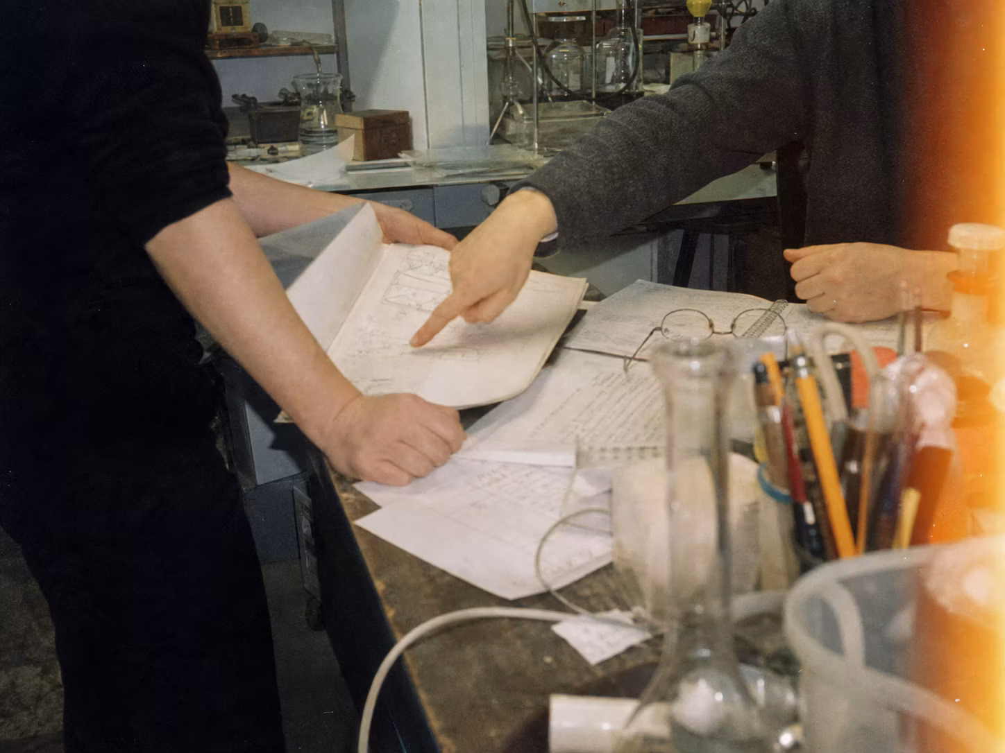Grainy amateur-film image of collaborators reviewing notes at a cluttered laboratory bench.