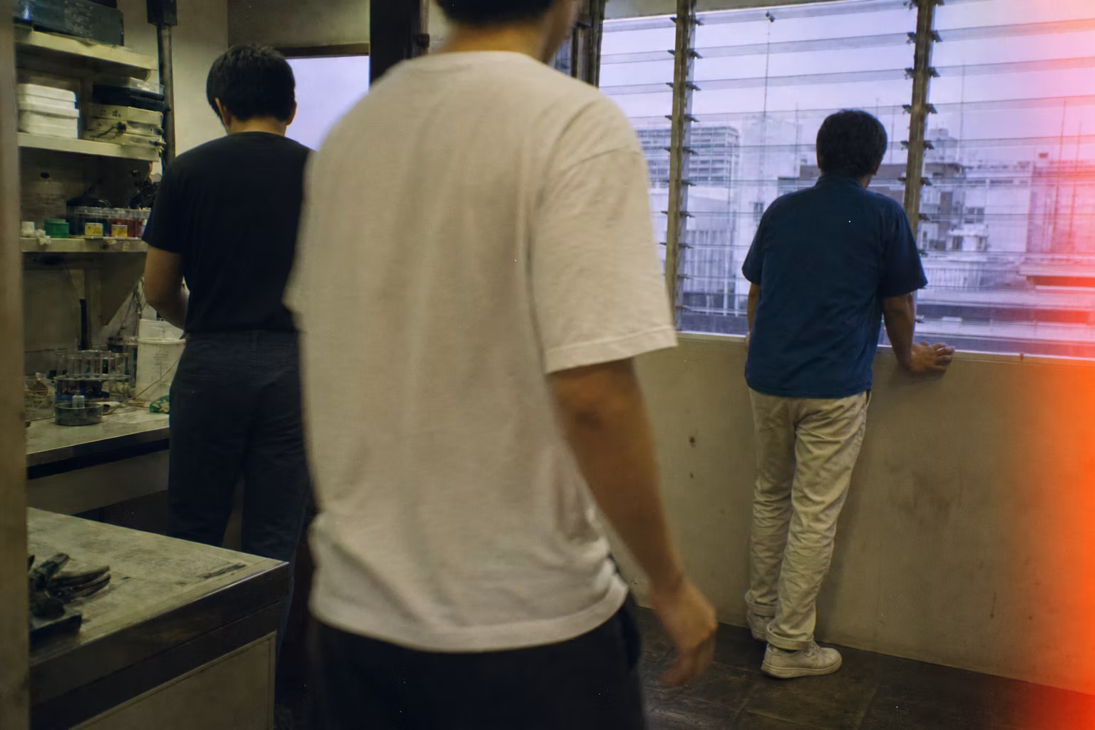 Grainy amateur-film image of anonymous collaborators moving through a Bangkok laboratory.