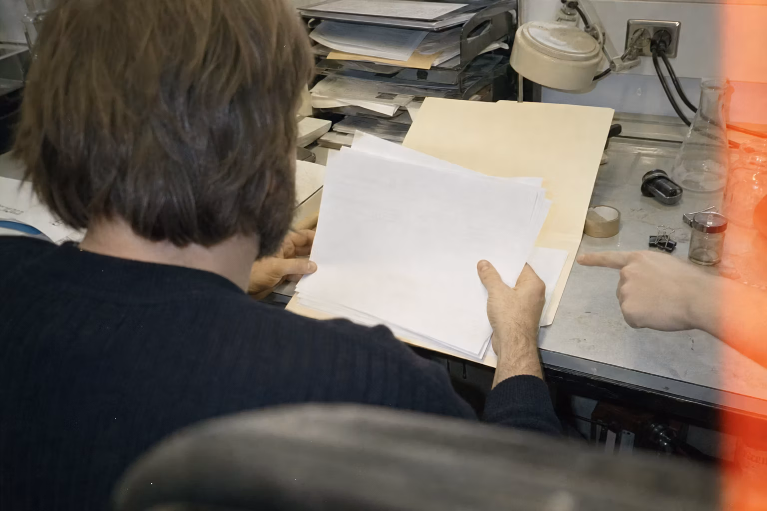 Grainy amateur-film image of collaborators reviewing unreadable documents at a cluttered laboratory desk.
