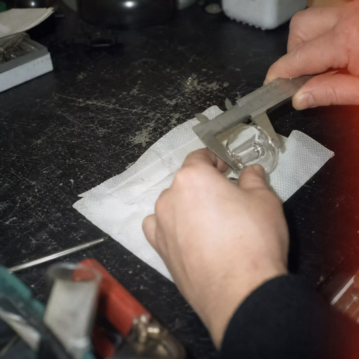 Grainy amateur-film image of hands making a precision check on an unlabelled glass component.